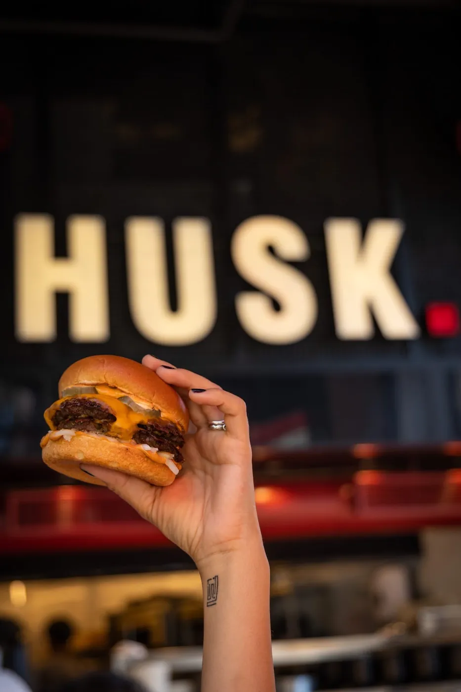 A hand holding up a Husk burger in front of the lit HUSK sign